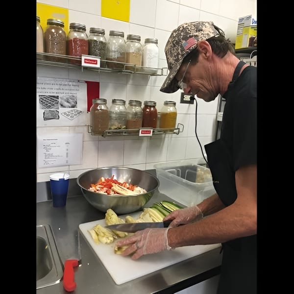 A deli worker in a camo hat and gloves chops vegetables on a cutting board.