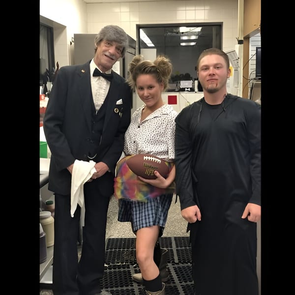 Three people in costumes, including a man in a suit, a woman with a football, and a man in a dark robe.