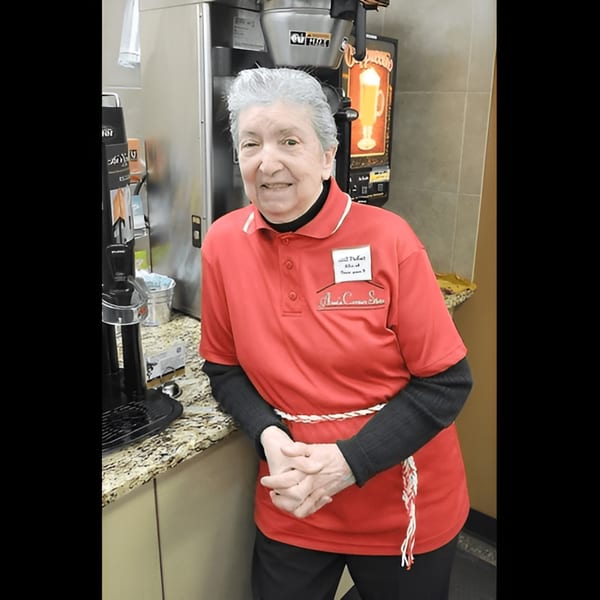 Smiling elderly woman in a red Ana's Corner Store shirt behind a deli counter.