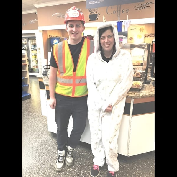 Two smiling people, one in a construction hat and safety vest, the other in a white onesie, inside Ana's Corner Store.