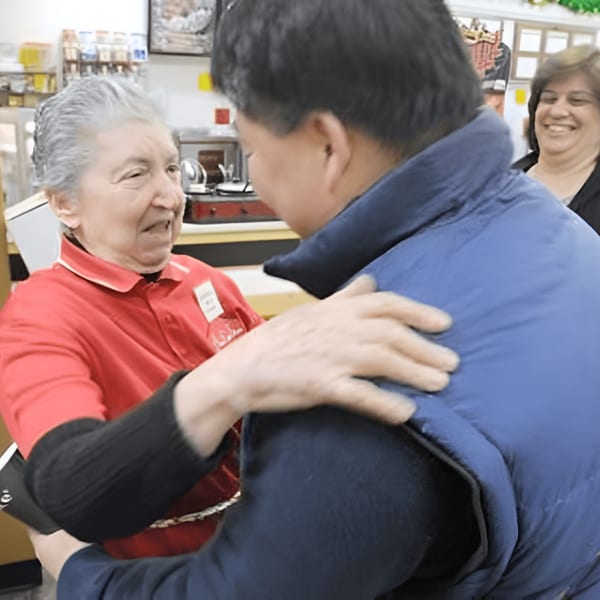 A smiling deli worker in a red shirt hugs a customer in a blue jacket at Ana's Corner Store.