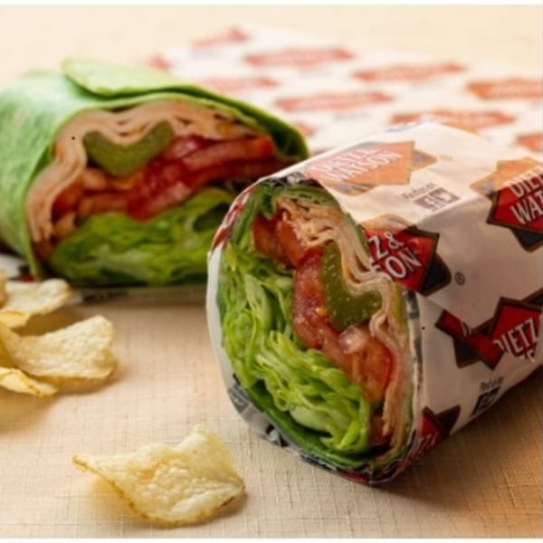 Two deli wraps, one in a green tortilla and one wrapped in paper, with chips on a table.