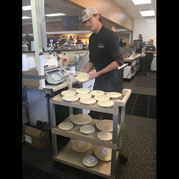 A deli worker in a cap and apron places a pie onto a cart filled with other pies.