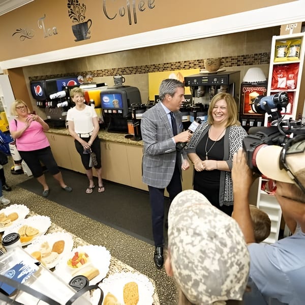 A reporter interviews a laughing woman in a deli, with other people and food displays in the background.