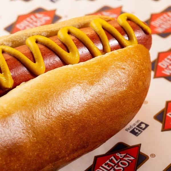 A hot dog with mustard in a bun, resting on branded deli paper from Ana's Corner Store.