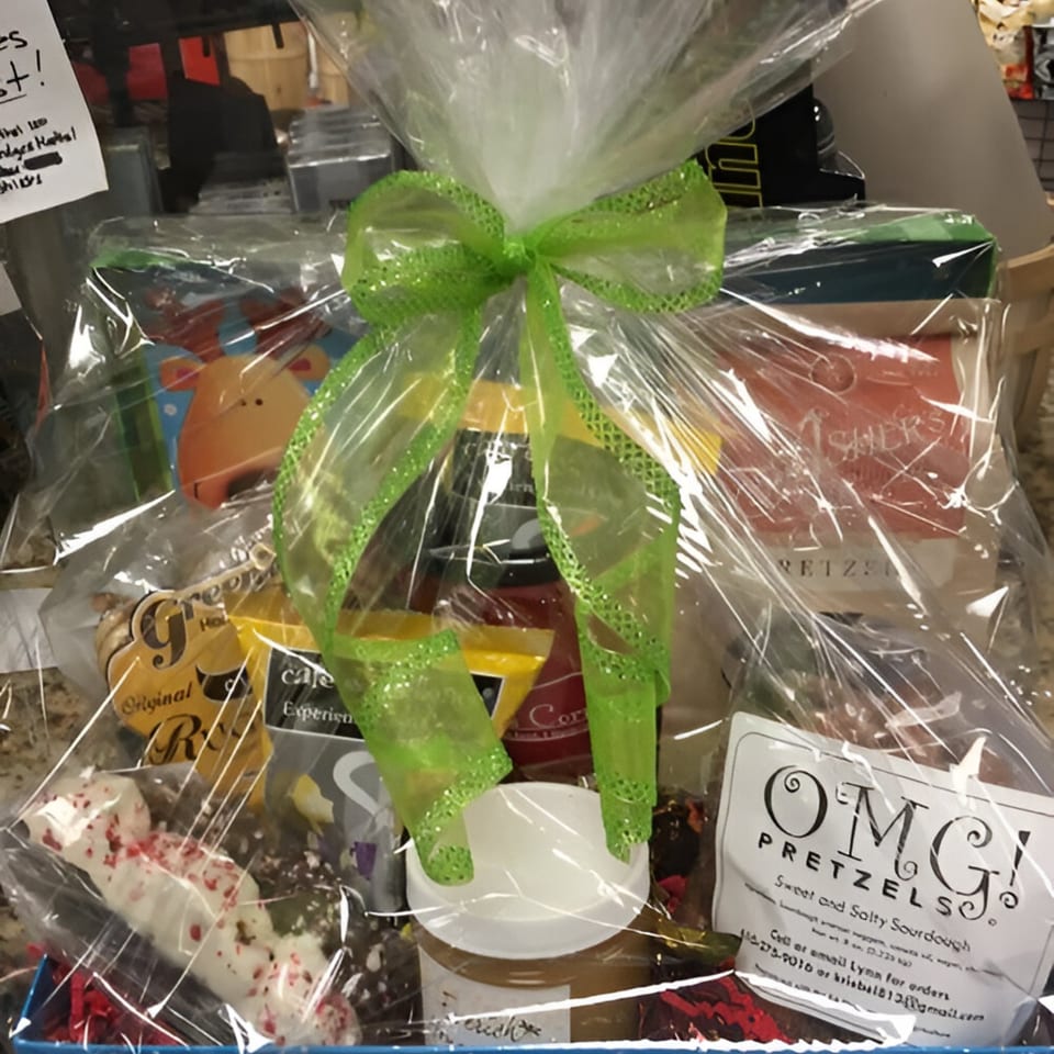 A deli gift basket filled with snacks, coffee, and pretzels, wrapped in cellophane with a green bow.