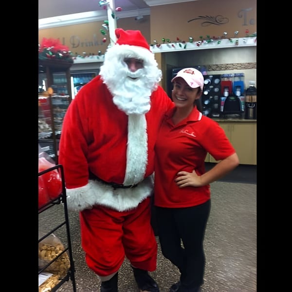 Santa Claus and a smiling Ana's Corner Store employee in a red shirt pose for a photo.