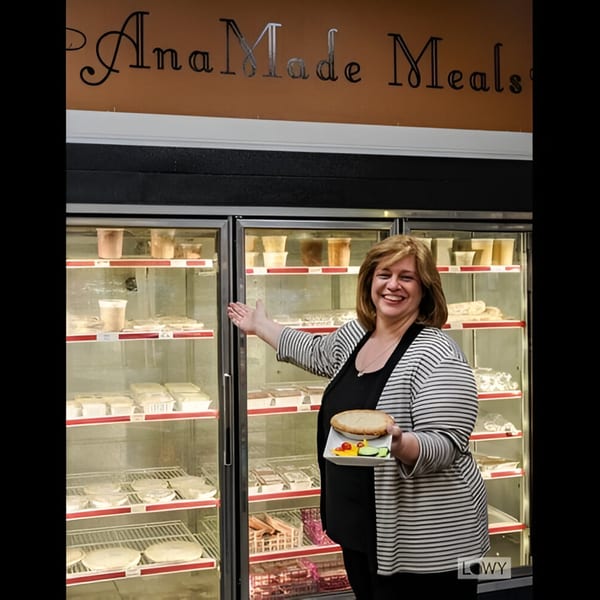 Smiling woman holding a pie and salad, gesturing to a deli display case filled with Ana's Corner Store meals.