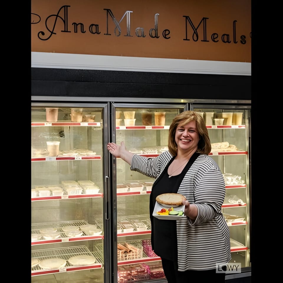 Smiling woman holding a pie and salad, gesturing to a deli display case filled with Ana's Corner Store meals.