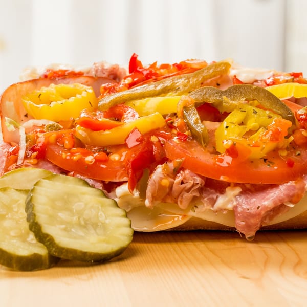Close-up of a deli sub sandwich with meats, cheese, tomatoes, and pickled peppers, with pickles on the side.