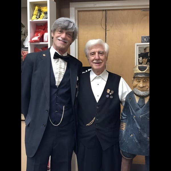 Two men in tuxedos and a wooden captain statue stand smiling in Ana's Corner Store.