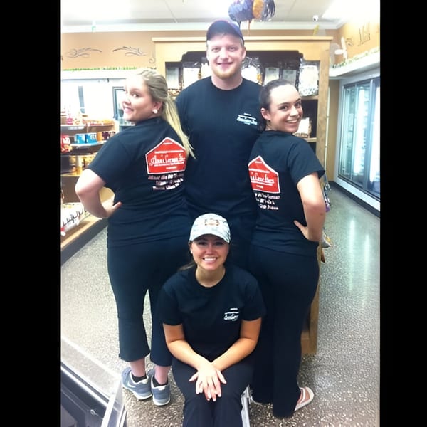 Four smiling Ana's Corner Store employees in black uniform shirts pose together inside the deli.