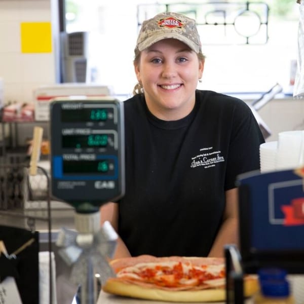Smiling deli worker in a Dietz & Watson hat behind a counter with a sandwich and scale.