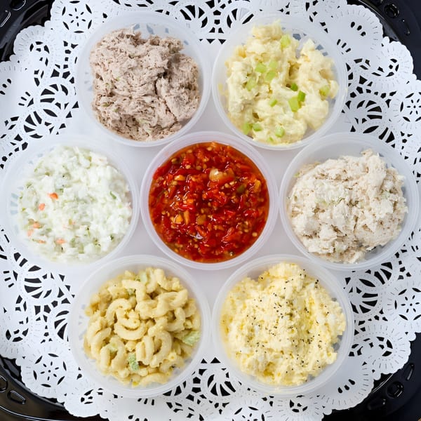 Seven deli salads in clear containers, including tuna, egg, macaroni, coleslaw, and a red relish.