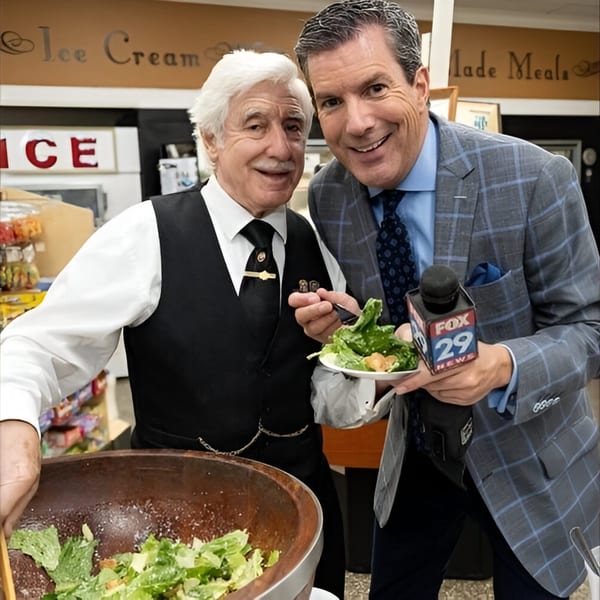 Two men, one in a vest and the other holding a FOX 29 News microphone, smile while serving salad.