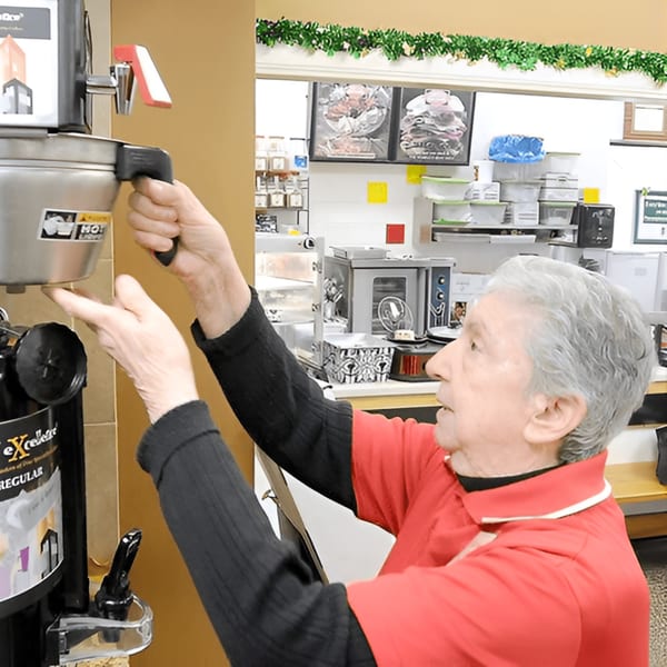 A deli worker in a red shirt operates a hot beverage dispenser at Ana's Corner Store.