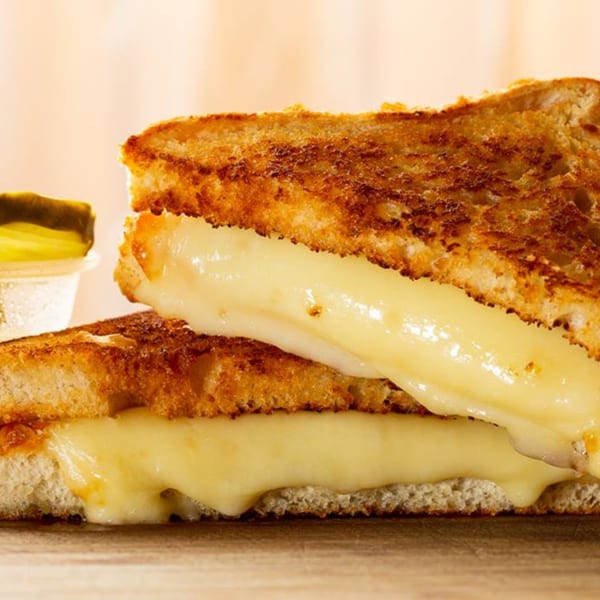 A grilled cheese sandwich from Ana's Corner Store, with melted cheese oozing out, and a side of pickles.
