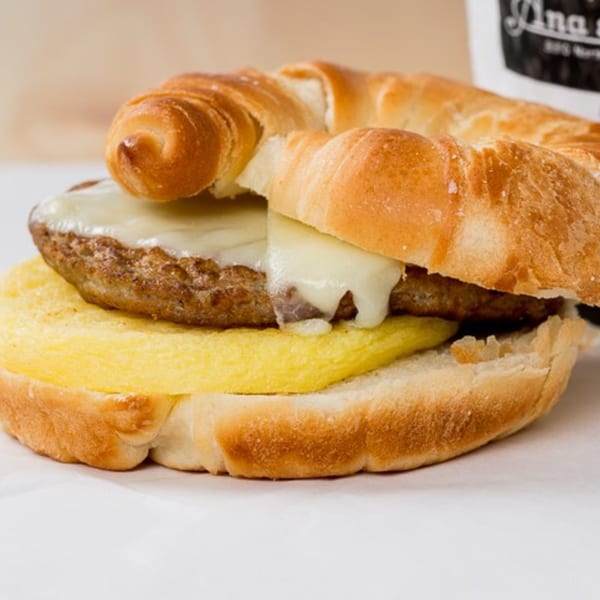 A breakfast sandwich from Ana's Corner Store with sausage, melted cheese, and egg on a croissant.