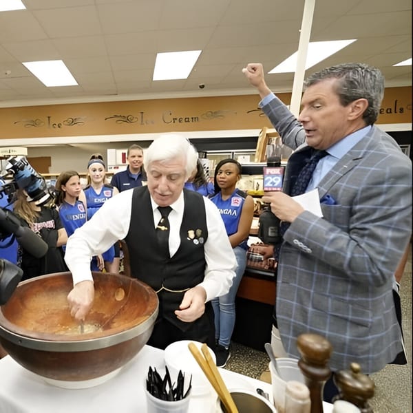 A man in a vest stirs a large bowl while a reporter with a microphone speaks beside him.