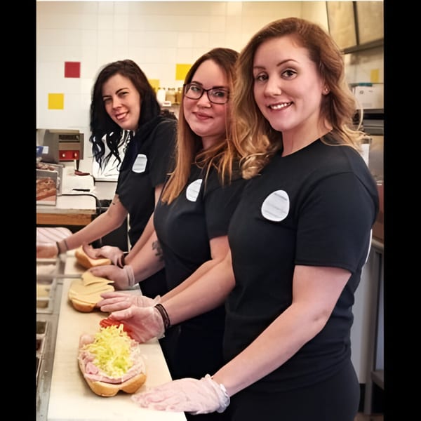 Three smiling women in black shirts and gloves prepare deli sandwiches at Ana's Corner Store.