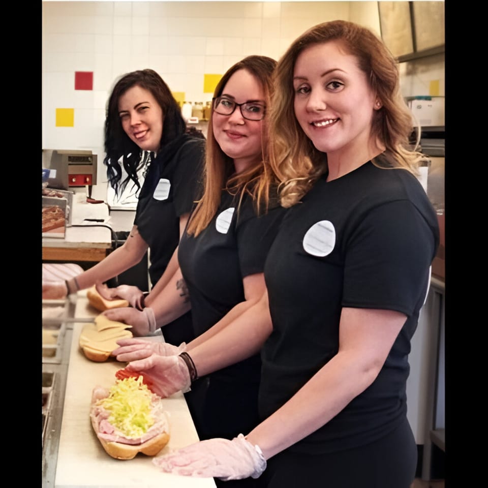 Three smiling women in black shirts and gloves prepare deli sandwiches at Ana's Corner Store.