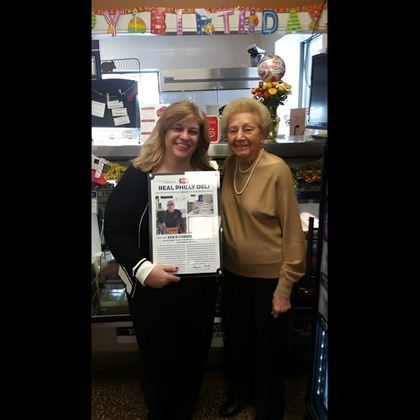 Two women smiling, one holding a framed article, in a deli with a "Happy Birthday" banner.