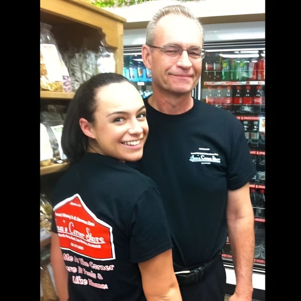 Two smiling Ana's Corner Store employees in black t-shirts stand in front of deli shelves.
