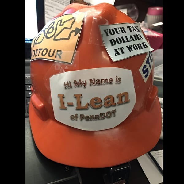 Orange hard hat with "Hi My Name is I-Lean of PennDOT" and other stickers.