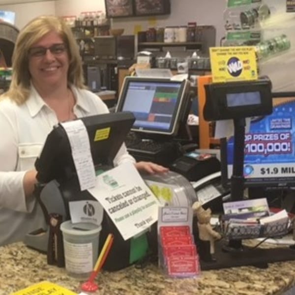 Smiling woman behind the counter at Ana's Corner Store deli, surrounded by registers and lottery signs.